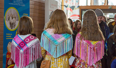Kolme saamelaista lasta seisoo selkä kuvaajaan päin harteillaan värikkäät huivit.
Kuvaaja: Eeva Mäkinen, Sámediggi/Saamelaiskäräjät