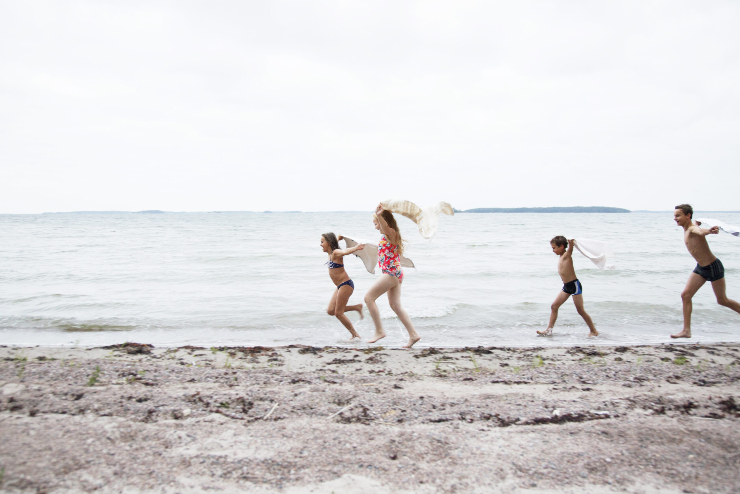 Valokuvaaja: Katri Lehtola Suomi-kuvapankki Lapset juoksevat rannalla uima-asuissa pyyhkeet hulmuten.